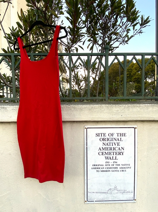 A red sleeveless dress on a black clothes hanger hanging from a tree at the Native American Cemetery Wall adjacent to Mission Santa Cruz