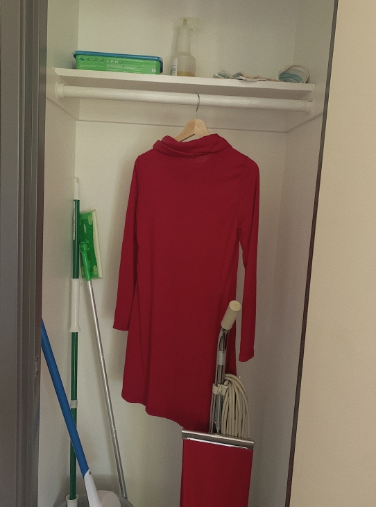 A long-sleeved red dress with a turtleneck hanging in a cleaning supply closet with a vacuum cleaner, broom, and mop